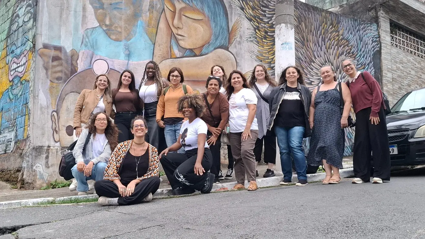 Grupo de mulheres reunido diante de mural com figuras femininas e grandes asas pintadas na Favela Galeria.