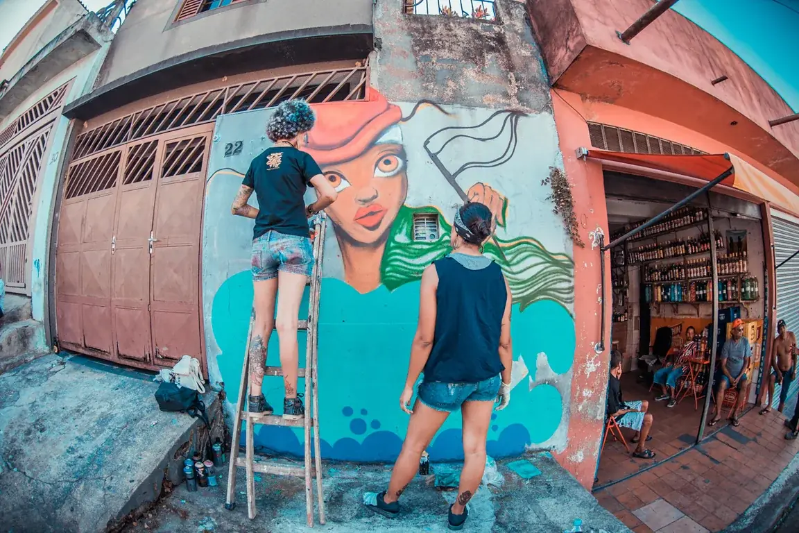 Mulheres pintando um grande mural com um rosto feminino na Vila Flávia, em registro da Ocupação Femi OPNI.