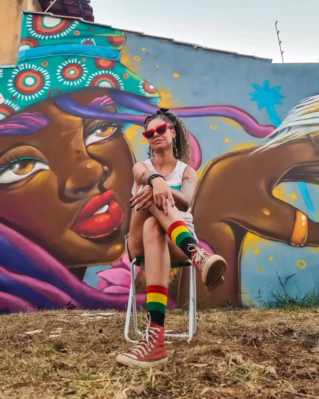 Crica Monteiro, de óculos vermelhos e meias listradas, sentada diante de um mural seu com um grande rosto feminino.