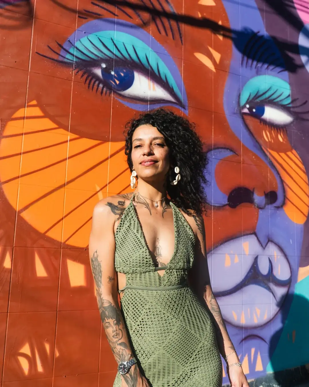 Laís da Lama, de vestido verde, diante de um painel seu com rostos femininos em laranja, lilás e azul.
