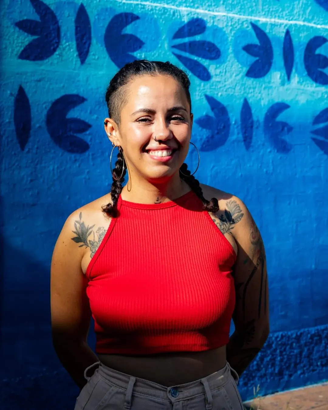 Nart sorri diante de uma parede azul pintada com folhas, de top vermelho e tranças.